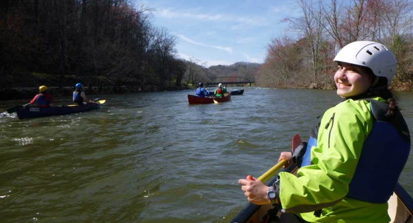 In the foreground, a person sitting at the front of canoe turns around and smiles for a photo. There are other canoes being navigated on the river in the background. 